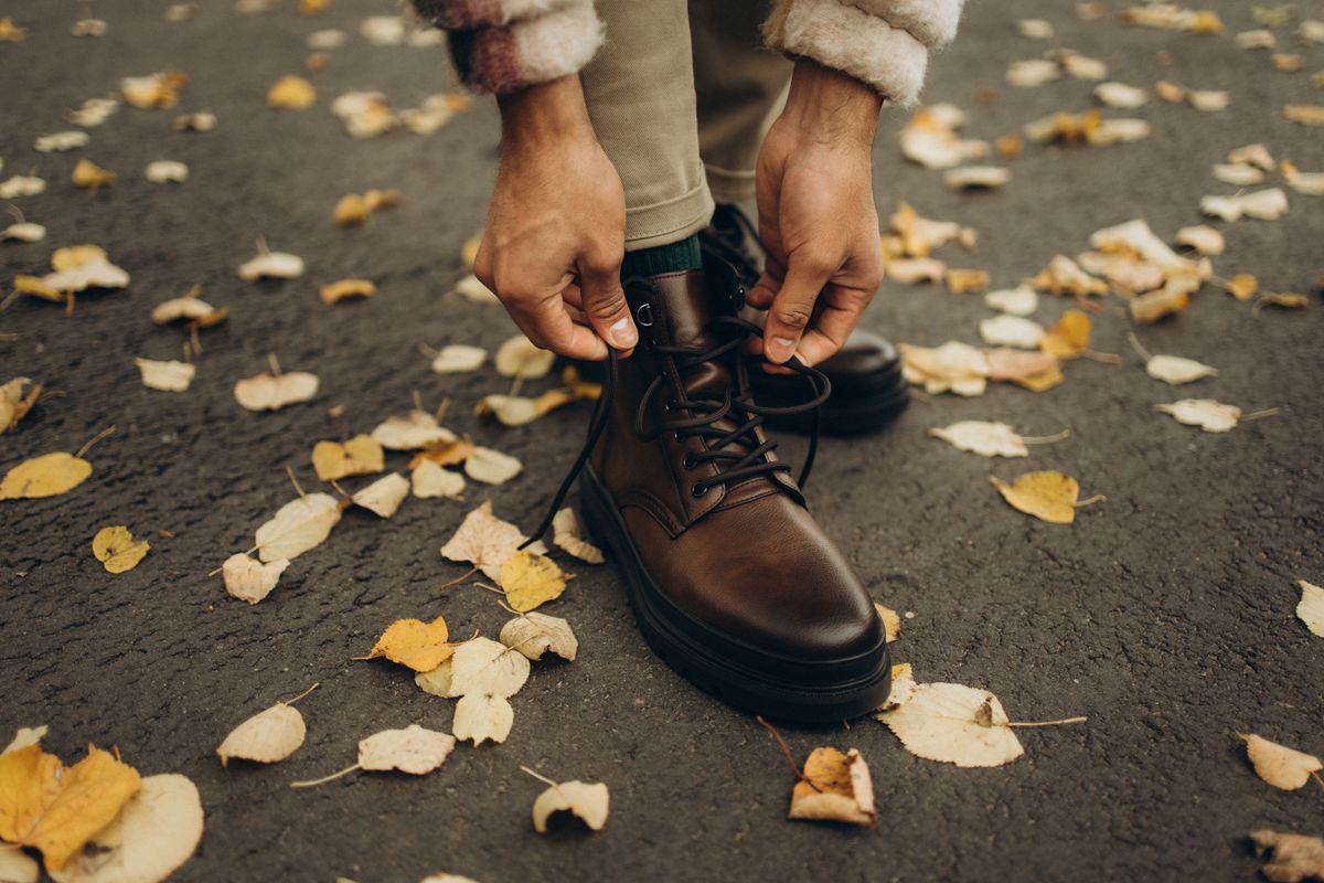Man lacing brown leather boots on a path covered in autumn leaves